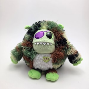 Może przedstawiać: Pluszowa zabawka potwora z kamuflażem w kolorze zielonym, brązowym i czarnym. Potwór ma zieloną twarz z fioletową opaską na oku, jednym dużym okiem i rzędem białych zębów. Zielone serce z napisem "PUSH" znajduje się na jego piersi.