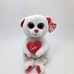 May include: A white plush bear with red accents on the ears, feet, and nose. The bear has large, sparkly pink and black eyes and holds a red heart embroidered with the word "love". A Ty Beanie Babies tag is attached.
