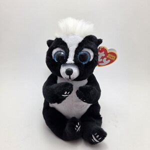 Peut inclure: Peluche de mouffette noire et blanche avec de grands yeux bleus et une touffe de cheveux blancs. La peluche a une étiquette rouge en forme de cœur attachée à l'oreille.