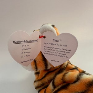 Op de afbeelding: Een pluche tijger Beanie Baby genaamd "India" met oranje en zwarte strepen. Het label is hartvormig en vermeldt "The Beanie Babies Collection" en "Geboortedatum: 26 mei 2000".