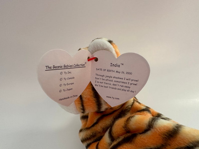 Op de afbeelding: Een pluche tijger Beanie Baby genaamd "India" met oranje en zwarte strepen. Het label is hartvormig en vermeldt "The Beanie Babies Collection" en "Geboortedatum: 26 mei 2000".