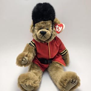 Può includere: Un orsacchiotto di peluche con pelliccia marrone chiaro che indossa una tunica rossa con finiture dorate e una cintura nera. L'orso indossa un alto cappello nero soffice e ha un'etichetta rossa a forma di cuore con le lettere ty sull'orecchio.