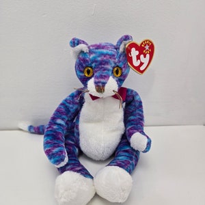 May include: Purple and blue striped plush cat with white fur on its belly and paws. The cat has yellow eyes and a red bow tie. The cat is sitting on a white surface. The tag reads "Beanie Baby" and "ty".