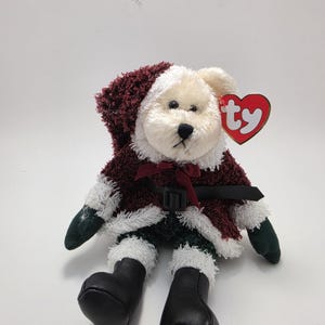 Può includere: Un orsacchiotto color crema che indossa un cappotto bordeaux testurizzato con cappuccio e bordo in pelliccia sintetica bianca, un sottile nastro bordeaux, una cintura nera con fibbia, muffole verde scuro e stivali neri. Un'etichetta Ty rossa.