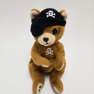 Op de afbeelding: Een bruine pluche teddybeer die een zwarte piratenhoed draagt met een wit schedel- en kruisbeenderontwerp. De beer heeft één oog bedekt met een zwart ooglapje.