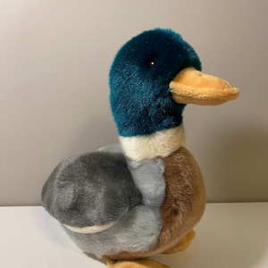 May include: A plush duck toy with a teal head, tan beak, and a mix of gray, brown, and blue-gray feathers. The stuffed animal has tan feet and a white collar.