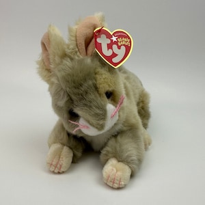 May include: A gray and white plush bunny with pink ears and paws. The bunny is lying down with its head turned to the side. The bunny has a Ty Beanie Babies tag attached to its ear.