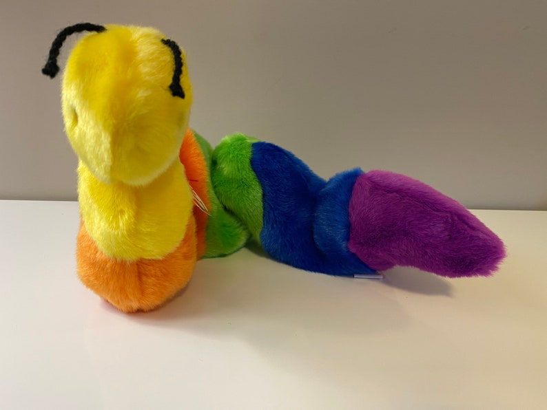 Op de afbeelding: Een pluche regenboogkleurige rups speelgoed met een gele kop en zwarte ogen. De rups is gemaakt van zacht, pluche stof en is ongeveer 30 cm lang.