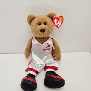 May include: A brown and white plush bear wearing a white basketball jersey with red and yellow trim. The jersey has the word "SHAQ" printed on the front. The bear is wearing black and red sneakers and has a red Ty Beanie Babies tag attached to its ear.