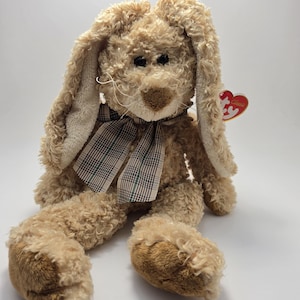 May include: A soft, tan-coloured bunny with long, floppy ears and a plaid ribbon around its neck. The bunny has a brown nose and dark eyes. A red heart-shaped tag is attached to the ear. The stuffed animal is a classic toy.