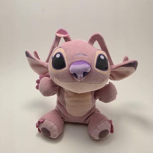 K&ouml;nnte beinhalten: Eine rosa Angel-Pl&uuml;schpuppe mit gro&szlig;en schwarzen Augen, einer lila Nase und gro&szlig;en Ohren. Die Puppe sitzt mit ausgestreckten Armen. Der K&ouml;rper der Puppe ist heller rosa als ihre Ohren und F&uuml;&szlig;e.