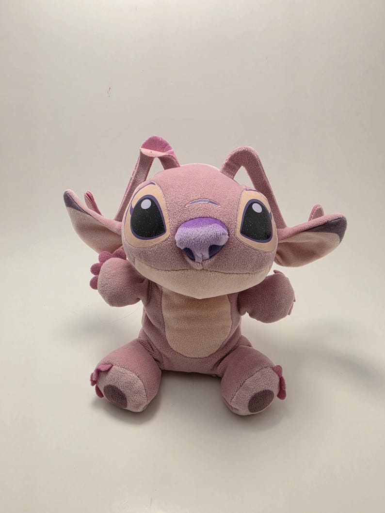 K&ouml;nnte beinhalten: Eine rosa Angel-Pl&uuml;schpuppe mit gro&szlig;en schwarzen Augen, einer lila Nase und gro&szlig;en Ohren. Die Puppe sitzt mit ausgestreckten Armen. Der K&ouml;rper der Puppe ist heller rosa als ihre Ohren und F&uuml;&szlig;e.