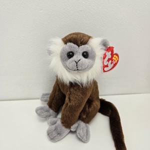 May include: A soft toy monkey with brown fur, grey hands and feet, and a white and grey face. It has a long brown tail and a red tag attached to its ear.