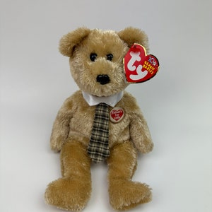 May include: A brown teddy bear wearing a white collar and a plaid tie. The bear has a red heart-shaped tag that says "Father's Day 2003" and a red tag that says "10th Beanie Babies".