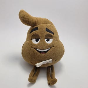 Op de afbeelding: Bruin pluche speelgoed in de vorm van een lachende poep emoji, met witte ogen en een klein, opgerold papieren accessoire. Het pluche speelgoed heeft een grillig ontwerp.