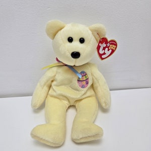 May include: A yellow plush Easter bunny beanie baby with a blue ribbon and an embroidered Easter egg on its chest. The tag reads "Ty Original Beanie Babies".