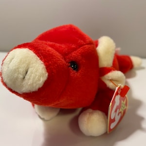 Puede incluir: Juguete de peluche rojo y blanco en forma de cerdo con una etiqueta Ty Beanie Baby. El cerdo está acostado de lado con la cabeza girada.