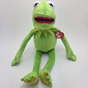 Può includere: Rana di peluche verde Kermit la rana seduta con le gambe incrociate. Ha un grande sorriso e un colletto giallo. Ha un'etichetta rossa Ty Beanie Baby attaccata al braccio.