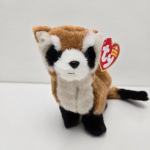 May include: A brown and white stuffed ferret with black feet and a black tail. The ferret is sitting on a white surface and has a red Ty Beanie Babies tag attached to its ear.