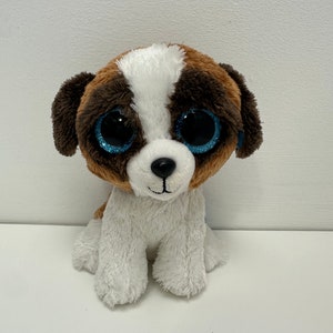 May include: A brown and white plush dog with large blue eyes. The dog is sitting with its paws tucked under its body.