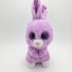 May include: A plush lavender bunny with large, glittery purple eyes and ears. The stuffed animal has a white muzzle and pink nose. The bunny's paws have white bottoms with bunny faces. A cute, cuddly toy.