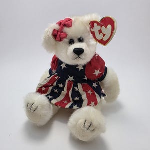 Può includere: Un orsacchiotto bianco che indossa un vestito con un motivo a stelle e strisce rosse, bianche e blu. L'orso ha un fiocco rosso e un'etichetta a forma di cuore. È un oggetto da collezione.