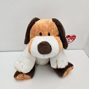 Puede incluir: Un peluche de perro con pelo marrón, beige y blanco. El perro tiene orejas caídas, un hocico blanco y ojos negros. Una etiqueta roja en forma de corazón está unida al juguete.