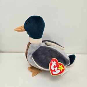 Pu&ograve; includere: Un'anatra di peluche grigia e blu con un becco marrone e zampe arancioni. L'anatra &egrave; seduta su una superficie bianca e ha un'etichetta rossa Ty Beanie Baby attaccata.