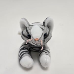 以下が含まれることがあります： グレーと白の縞模様、白い足、青い目の白い顔をしたぬいぐるみ猫。猫はピンクのひげとピンクの鼻をしています。おもちゃは白い面に横たわっています。