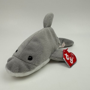 Puede incluir: Juguete de peluche de delfín gris con ojos negros y vientre blanco. El juguete tiene una etiqueta TY roja adjunta.