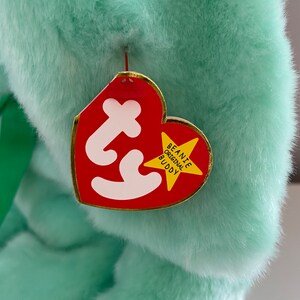 Puede incluir: Un peluche verde menta con una etiqueta roja en forma de coraz&oacute;n. La etiqueta tiene la palabra "Ty" en blanco y una estrella con borde dorado que dice "Beanie Original Buddy". Se ve una cinta verde.