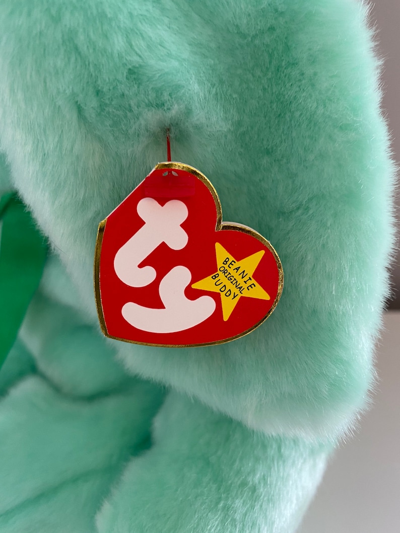 Puede incluir: Un peluche verde menta con una etiqueta roja en forma de coraz&oacute;n. La etiqueta tiene la palabra "Ty" en blanco y una estrella con borde dorado que dice "Beanie Original Buddy". Se ve una cinta verde.