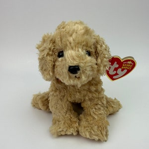 May include: A plush, tan-colored dog toy with a curly fur texture. The toy has black eyes and a black nose. A red heart-shaped tag with white text is attached to the dog's ear. The dog is sitting on a white surface.
