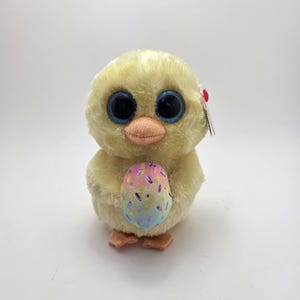 May include: A plush yellow chick with large blue eyes and a peach-colored beak. The chick is holding a colorful Easter egg with pastel sprinkles. A small red tag is attached to the chick's ear. The toy is designed for children.