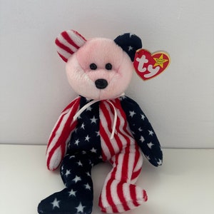 May include: A pink and white beanie baby bear wearing a red, white, and blue American flag pattern. The bear has black eyes and a black nose. The bear is sitting on a white surface. The bear has a red heart-shaped tag that says "Beanie Baby" on it.