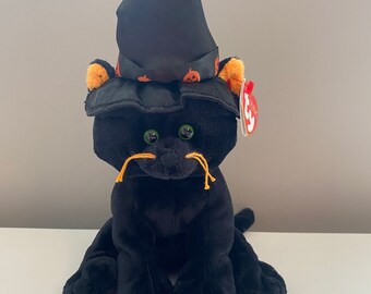 Puede incluir: Un juguete de peluche negro con forma de gato que lleva un sombrero de bruja negro con detalles de calabaza naranja. El gato tiene ojos verdes, detalles de orejas naranjas y bigotes naranjas. Una etiqueta roja está adjunta al gato.
