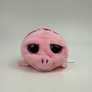 May include: A plush pink turtle toy with large, glittery purple-rimmed eyes. The turtle has a light pink body with darker pink accents on its head and a small, smiling mouth. A small tag is attached.