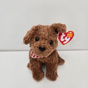 May include: A brown plush puppy with a red and white checkered bandana around its neck. The stuffed animal has a black nose and large, round eyes. A red heart-shaped tag is attached to the ear.