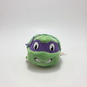 Puede incluir: Un peluche verde de Donatello, una Tortuga Ninja. El juguete tiene una máscara morada, ojos blancos y una sonrisa cosida. El peluche es pequeño y redondo, con una etiqueta adjunta.