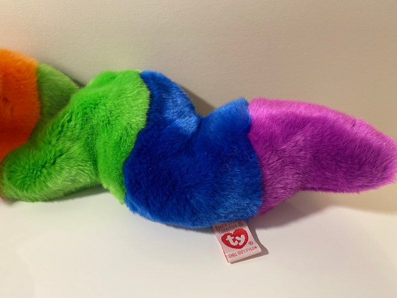 Op de afbeelding: Een veelkleurig pluche speelgoed in de vorm van een worm. De worm is oranje, groen, blauw en paars. Het speelgoed heeft een Ty-label eraan bevestigd met de tekst "Ty, Beanie Babies, Small  (6"  tall)".