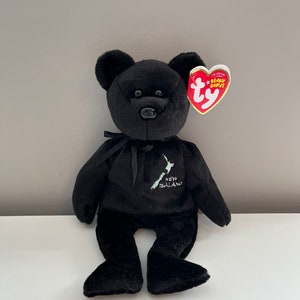 May include: Black plush beanie baby bear with a black ribbon around its neck. The bear has a white embroidered outline of New Zealand on its chest with the text "New Zealand". The bear has a red Ty Beanie Babies tag attached to its right ear.