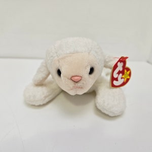 May include: A plush white lamb toy with a pink nose and black eyes. The lamb has a tag attached to its ear. The toy is lying down on a white surface.