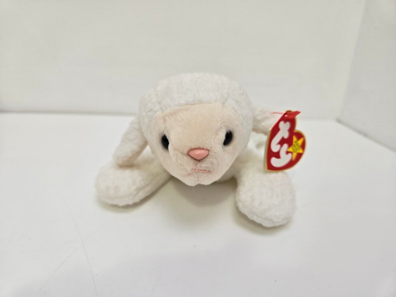 May include: A plush white lamb toy with a pink nose and black eyes. The lamb has a tag attached to its ear. The toy is lying down on a white surface.