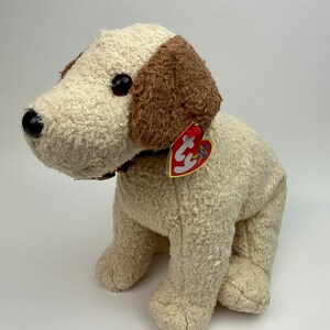 Op de afbeelding: Een wit en bruin pluche hondenspeelgoed met een rood hartvormig label waarop "Ty" staat. De hond zit met gekruiste poten en heeft een bruine halsband.