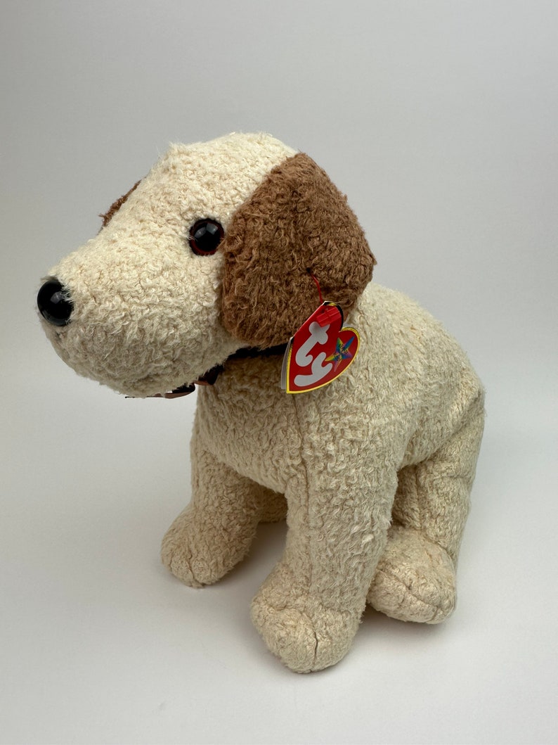 Op de afbeelding: Een wit en bruin pluche hondenspeelgoed met een rood hartvormig label waarop "Ty" staat. De hond zit met gekruiste poten en heeft een bruine halsband.