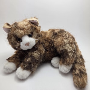 May include: A soft toy cat in brown and white. The cat has a fluffy, textured coat with darker brown markings and white paws and muzzle. The cat's ears are a pale pink colour.