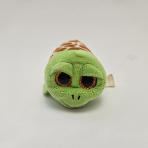 May include: A small plush turtle toy with a light green body and a brown and white patterned shell. The toy features large, round, glittery orange eyes and a soft, textured fabric surface. A small white fabric tag is visible on the side.