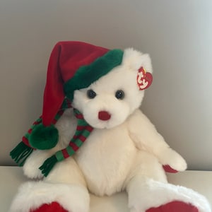 Może przedstawiać: Biały pluszowy miś w czerwono-zielonej, paskowanej szalce i czerwono-zielonej czapce Świętego Mikołaja. Miś ma czerwony nos i czarne oczy. Miś siedzi na białym tle.