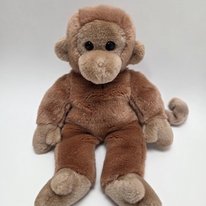 May include: A brown plush monkey toy with black eyes and a long tail. The monkey is sitting with its legs crossed and its arms at its sides.