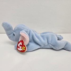 May include: A light blue plush elephant toy with a pink nose and ears. The toy is lying on its side with its head turned to the side. The toy has a red and yellow Ty tag attached to its leg.